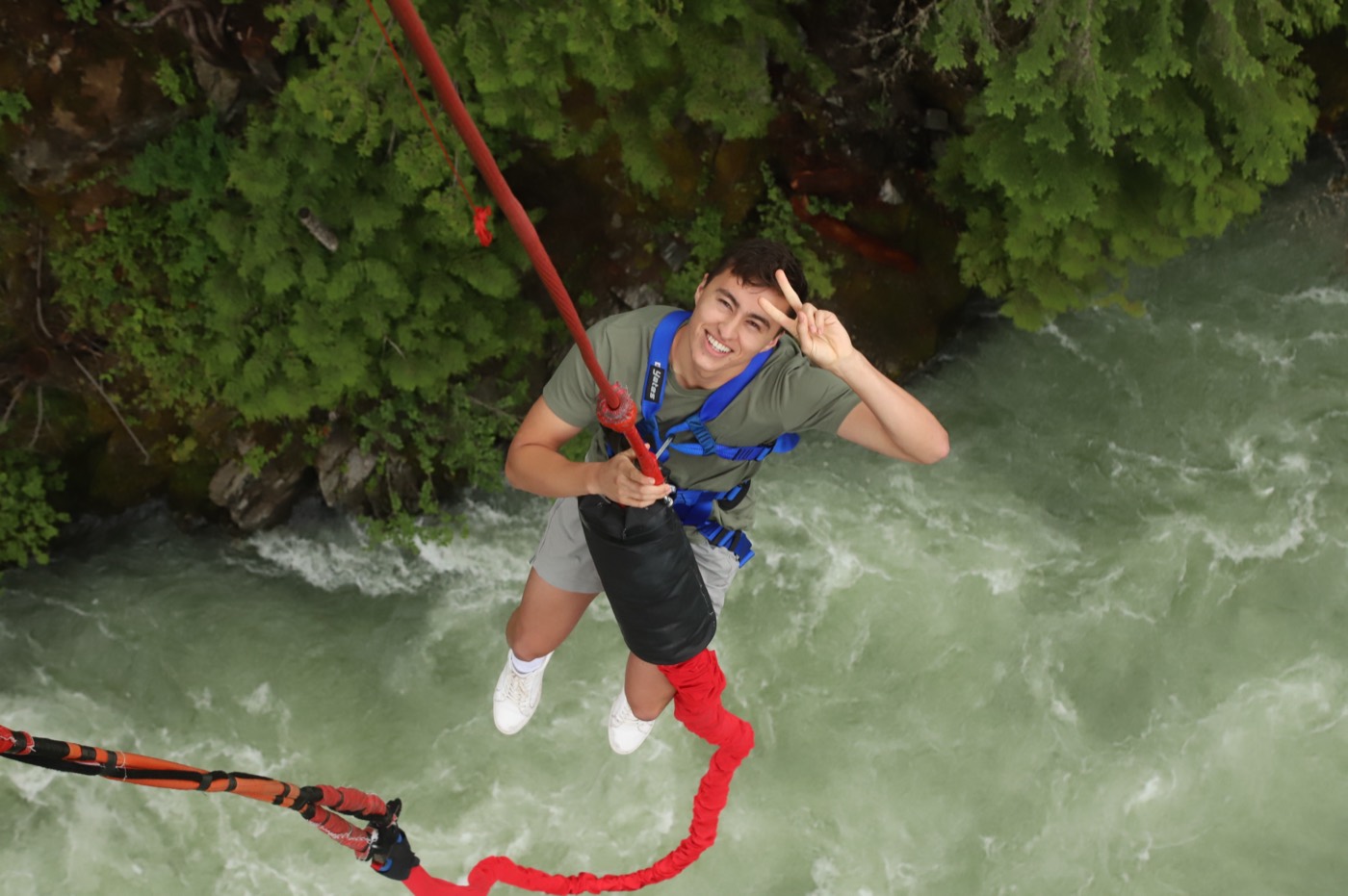 Cesar mid bungee jump above a rushing river, flashing a peace sign