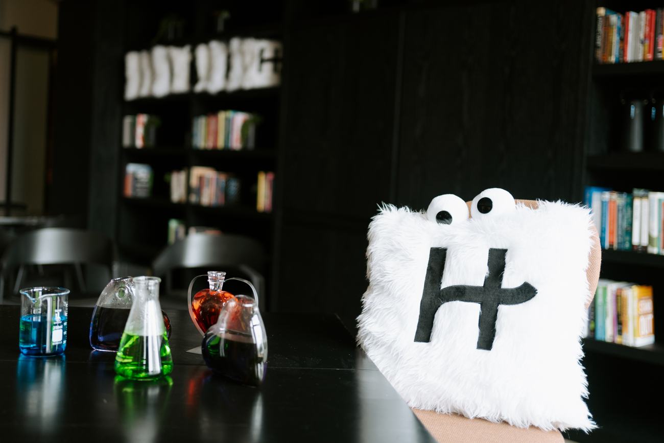 The fuzzy Hinge mascot pillow beside colorful beakers and flasks styled like a science lab